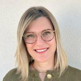 Porträt einer lächelnden Frau mit Brille, blondem Haar und grünem Oberteil vor neutralem Hintergrund.